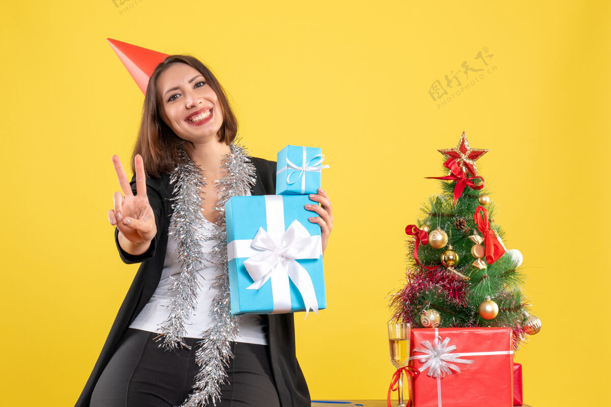 圣诞节圣诞气氛积极美丽的女士戴着圣诞帽拿着礼物在黄色的办公室里摆着胜利的手势举行礼物美丽的女士