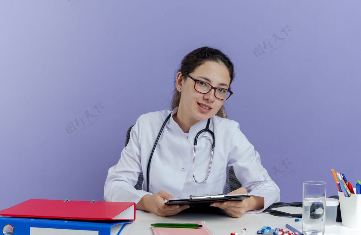 医生高兴的年轻女医生穿着医用长袍和听诊器坐在办公桌旁 带着医疗工具 看起来与世隔绝坐着剪贴板年轻人