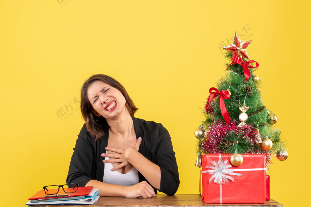 电脑情绪激动的年轻女子坐在一张桌子旁装饰圣诞树在黄色办公室树微笑圣诞