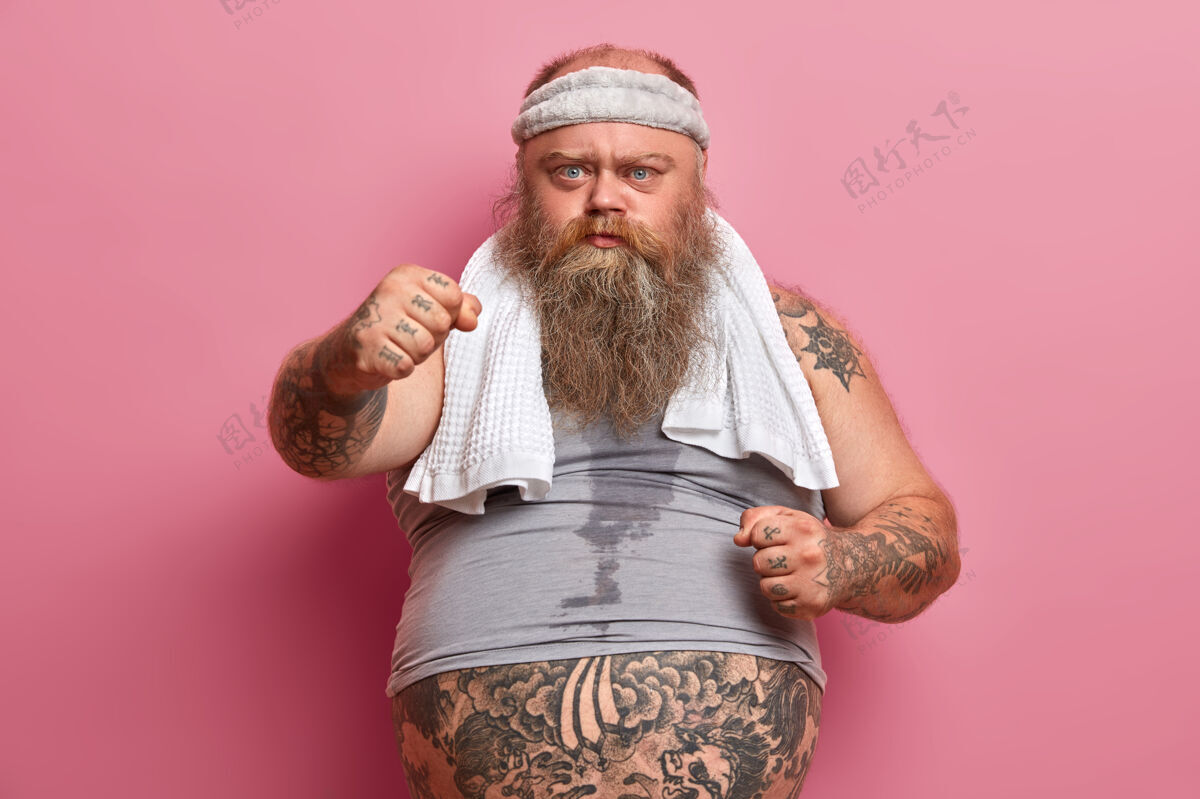 体位严肃的胖乎乎的胡须男人握紧拳头 忍受额外的体重 喜欢运动 出汗的身体和纹身的手臂 对着粉色的墙壁摆姿势减肥和节食的概念体重腹部站立