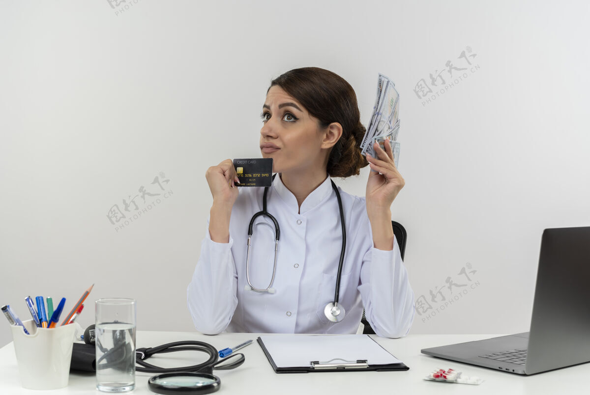 坐着体贴的年轻女医生穿着医用长袍和听诊器坐在办公桌旁 手里拿着医疗工具和笔记本电脑 手里拿着信用卡和钱 孤零零地抬头看听诊器医疗成人