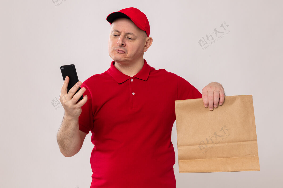 帽子一个穿着红色制服 戴着帽子的送货员拿着一个纸包 严肃地看着手机屏幕 站在白色的背景上严肃屏风包装