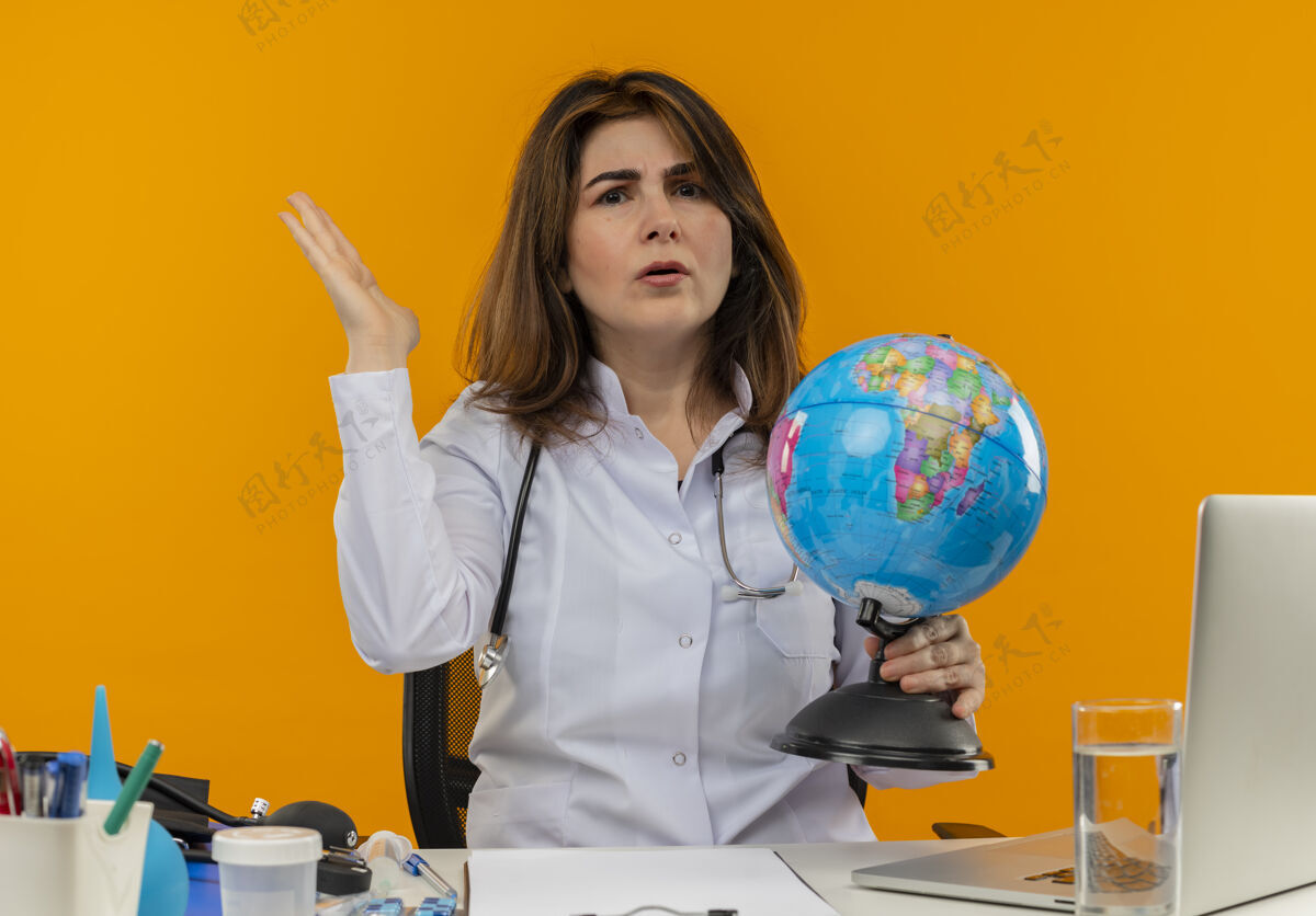 医疗未出租的中年女医生穿着医用长袍和听诊器坐在办公桌旁 手持医疗工具笔记本电脑和剪贴板拿着地球仪 显示空手孤立不愉快拿着穿