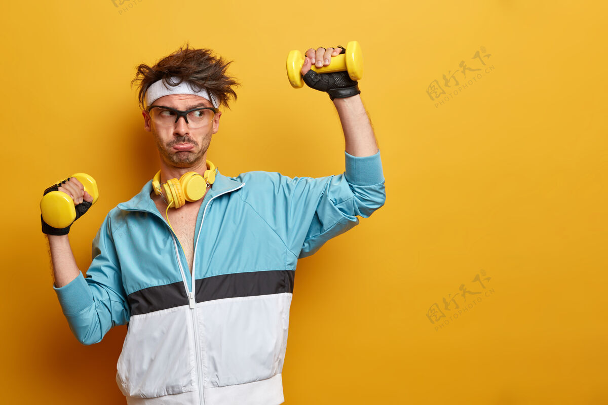 积极运动健将举起哑铃 努力训练肱二头肌 过着积极健康的生活方式 有规律的体育锻炼 靠着黄墙摆姿势 空荡荡的一边肌肉眼镜哑铃