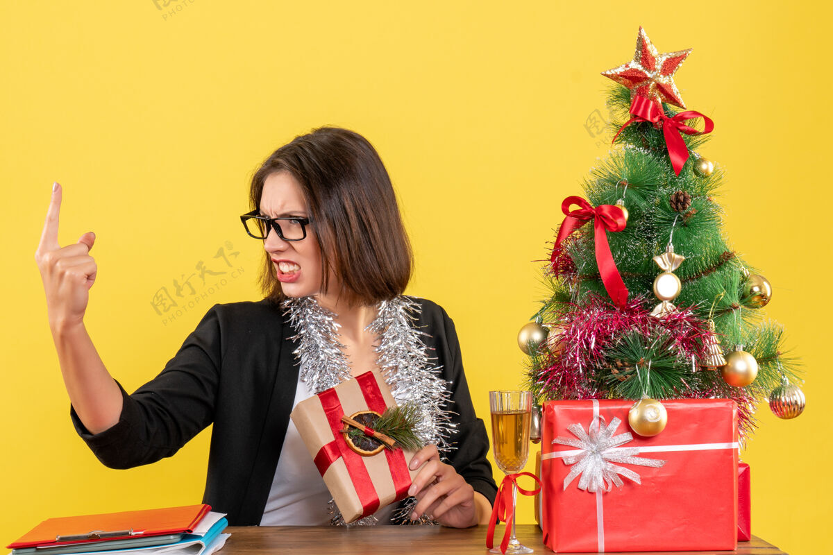 新郎一位穿着西装 戴着眼镜的漂亮商务女士怒气冲冲地朝上指 坐在一张桌子旁 桌子上有一棵黄色的圣诞树西装人人