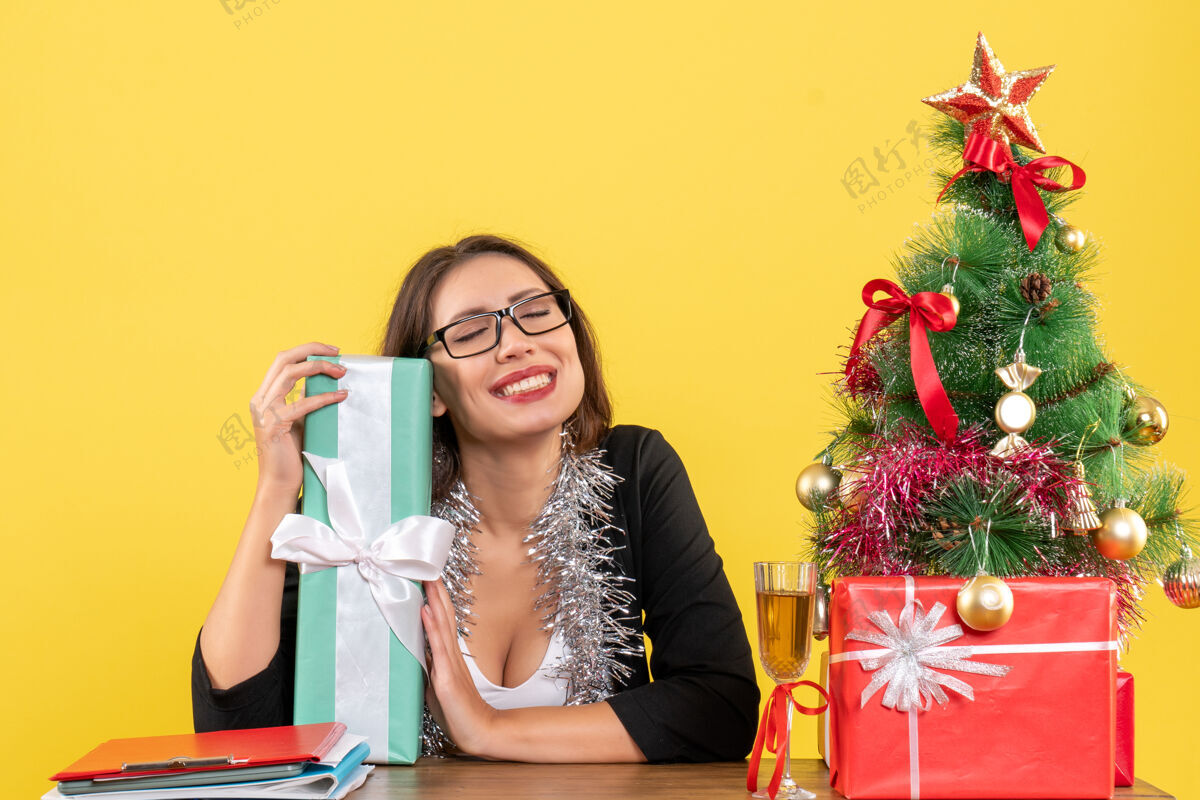 秀一位穿着西装 戴着眼镜的商务女士展示着她的礼物 她梦想着什么 坐在办公室的桌子旁 桌上放着一棵圣诞树成人眼镜桌子