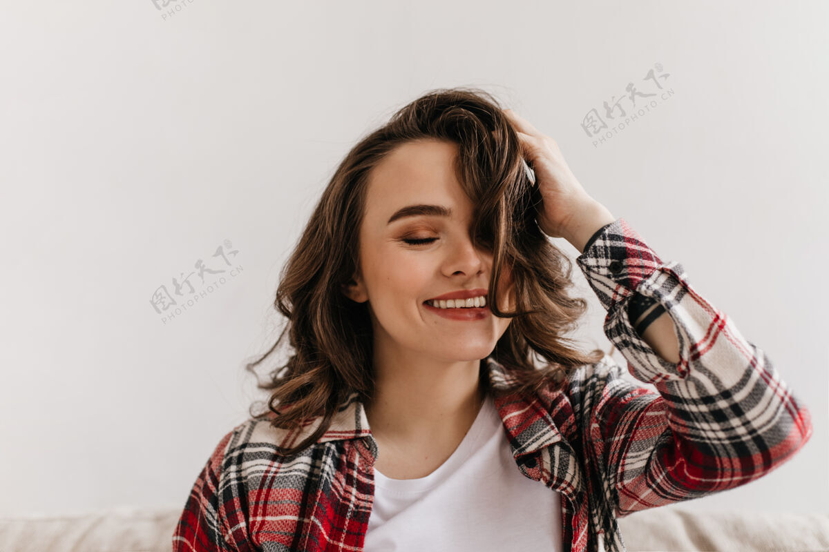 美丽穿着格子衬衫的快乐的年轻女子微笑着玩头发坐着房子期望
