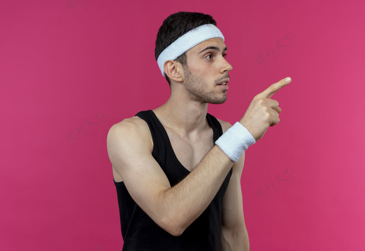 站立戴着头巾的年轻运动型男子一边看一边用手指指着粉色背景上的某个东西手指指尖头带
