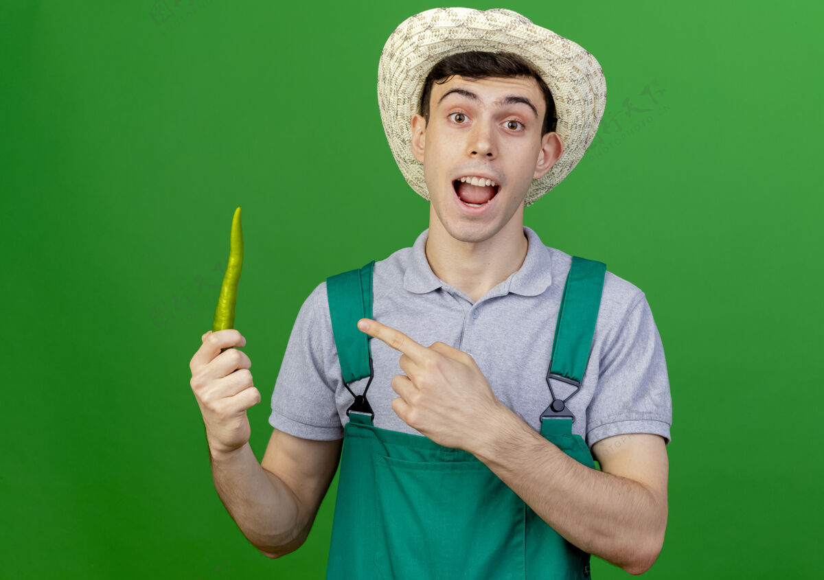 穿惊讶的年轻男园丁戴着园艺帽 指着隔离在绿色背景上的辣椒和复印空间胡椒点绿色