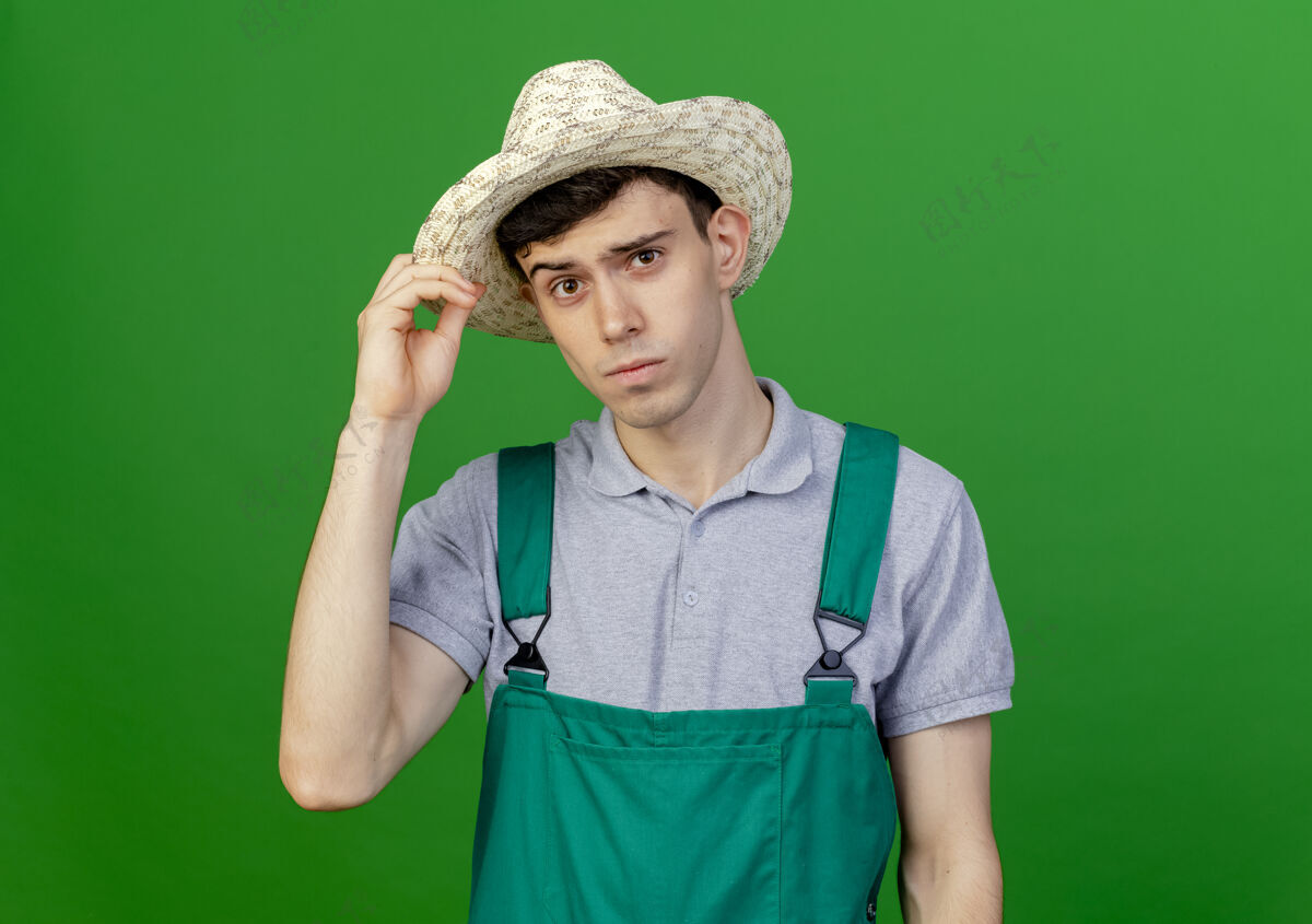 园艺自信的年轻男性园丁戴着和拿着园艺帽隔离在绿色背景与复制空间空间帽子复制