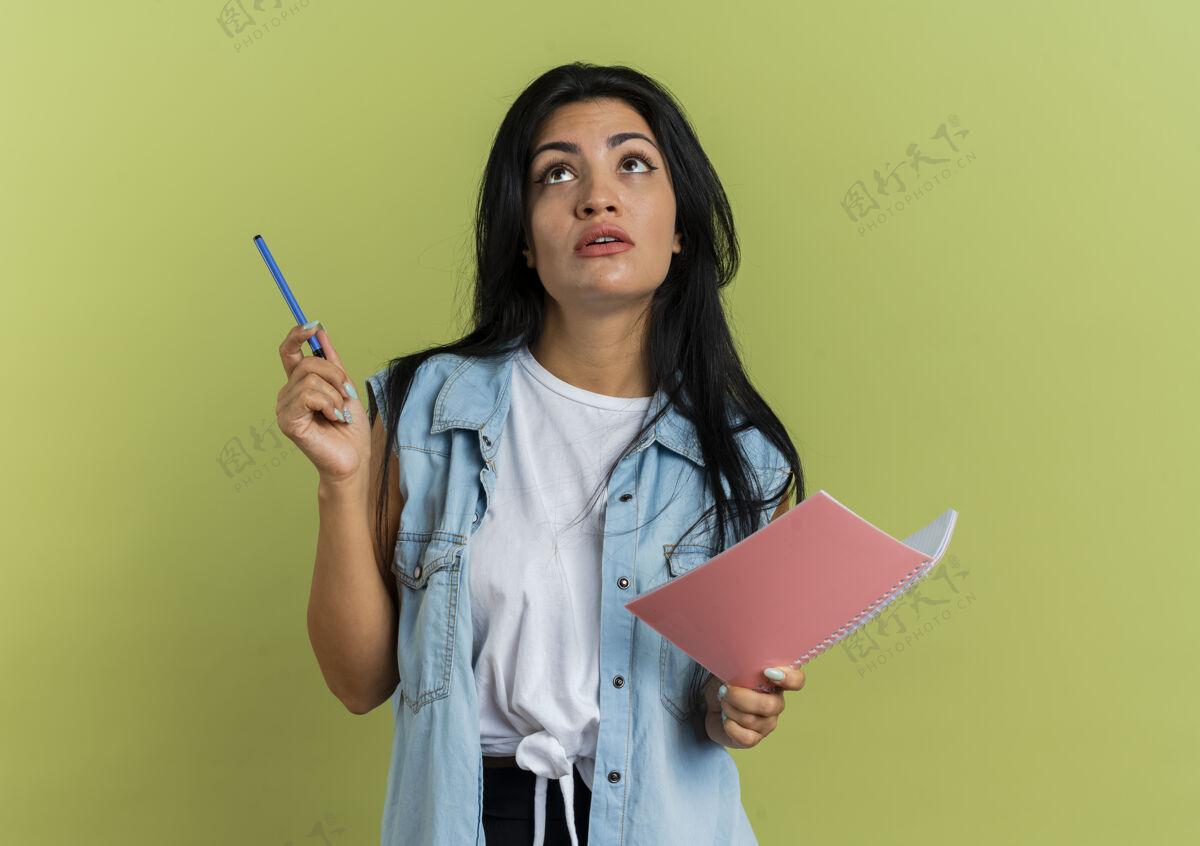 笔记本震惊的年轻白人女孩拿着钢笔和笔记本 在橄榄绿的背景和复印空间上孤立地抬头看空间持有绿色