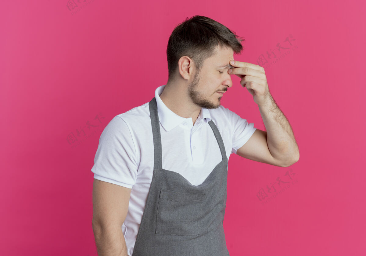 累了围着围裙的理发师看起来很累 而且工作过度 他闭着眼睛站在粉色背景上摸鼻子摸着理发师眼睛