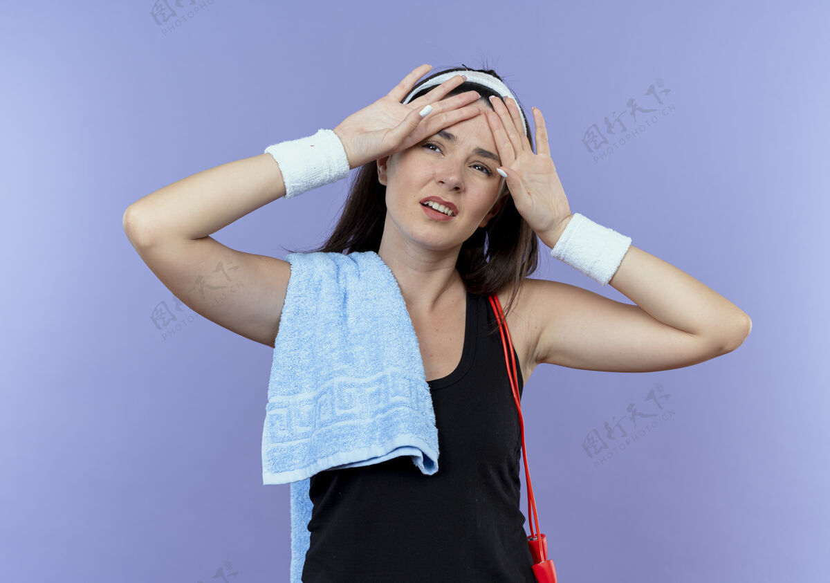 锻炼戴着头巾 肩上扛着毛巾的年轻健身女士 站在蓝色背景上锻炼后显得疲惫和疲惫筋疲力尽肩膀健身