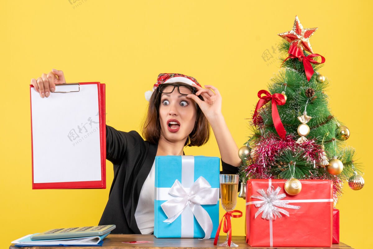 快乐迷人的女士穿着西装 戴着圣诞老人的帽子 在办公室里查看文件 感觉很惊讶 就在黄色隔离带上检查文件人