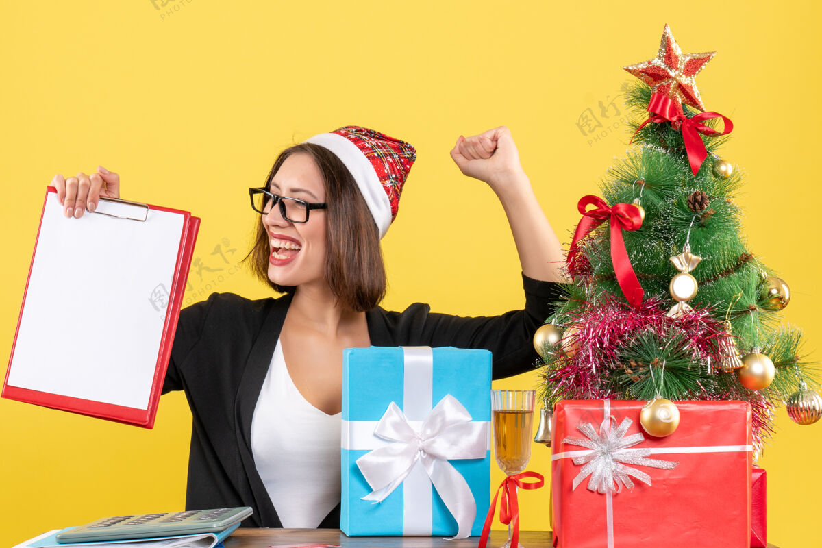 黄色迷人的女士穿着西装 戴着圣诞老人的帽子和眼镜 骄傲地拿着文件在办公室里文件持有漂亮