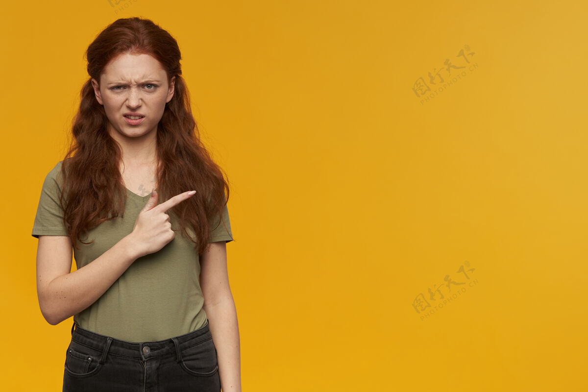 手指愤怒的 皱眉的女人 长着姜黄色的头发 穿着绿色的t恤 情感的概念食指指向右边的复制空间 隔离在橙色的墙上姜女士独自