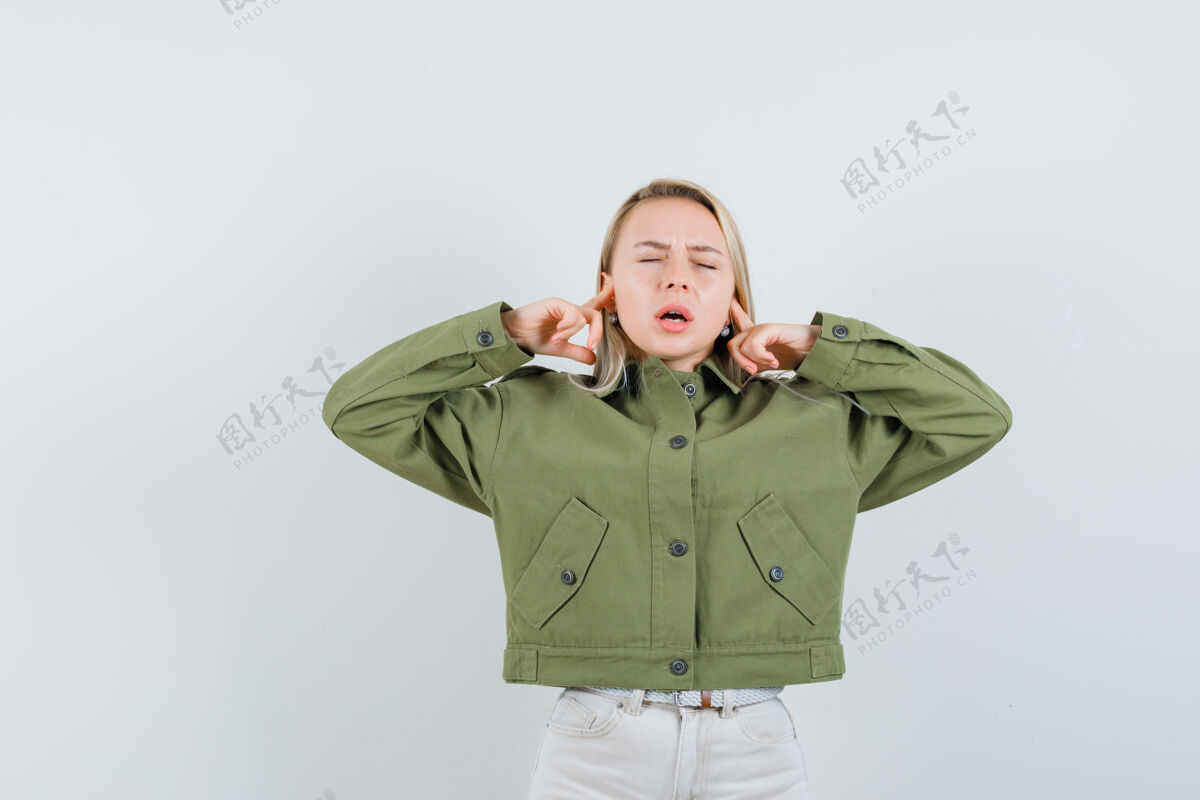 夹克穿着绿色夹克的年轻女性 牛仔裤用手指塞住耳朵 看起来很恼火 正面照健康成人皮肤