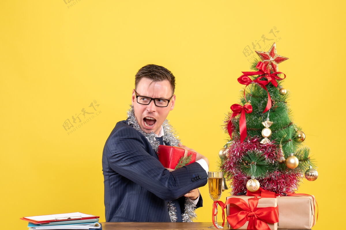人前视图激动的男人把礼物藏在圣诞树旁的桌子上 黄色背景上的礼物微笑坐着前面