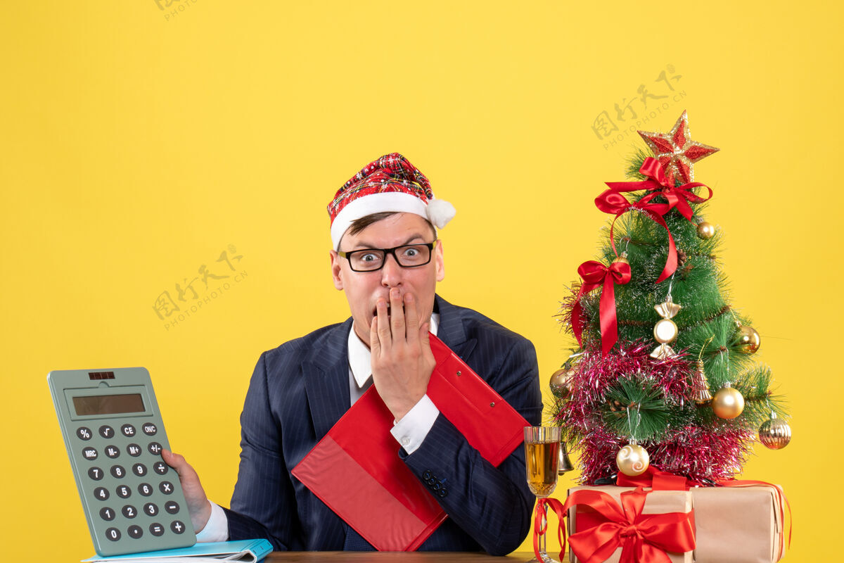 背景前视图激动的商人拿着计算器坐在圣诞树旁的桌子上 呈现黄色背景抱着坐着专业