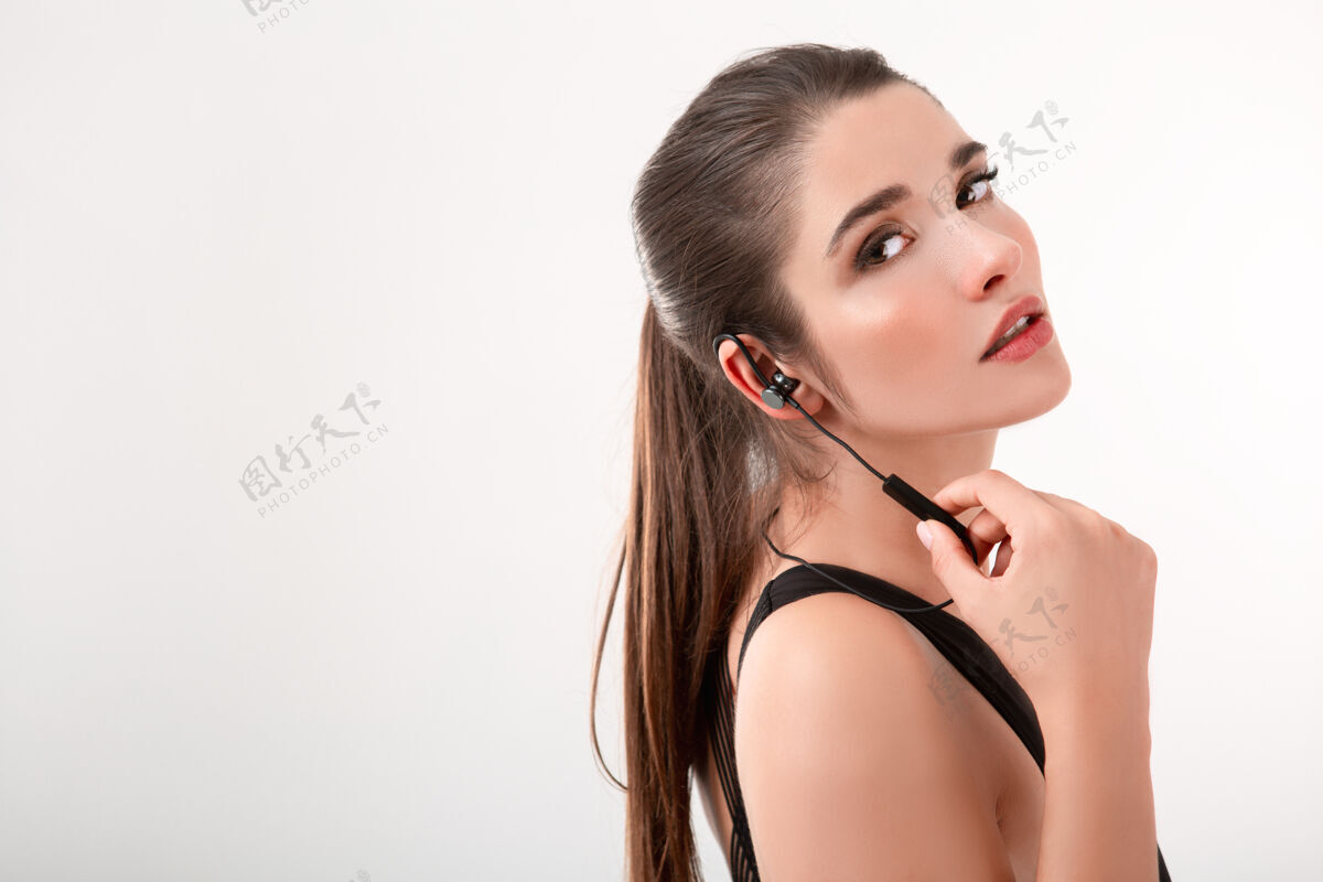 耳机迷人的黑发女子穿着黑色上衣慢跑 戴着耳机听音乐 在白色墙壁上摆出孤立的马尾发型运动员女孩慢跑