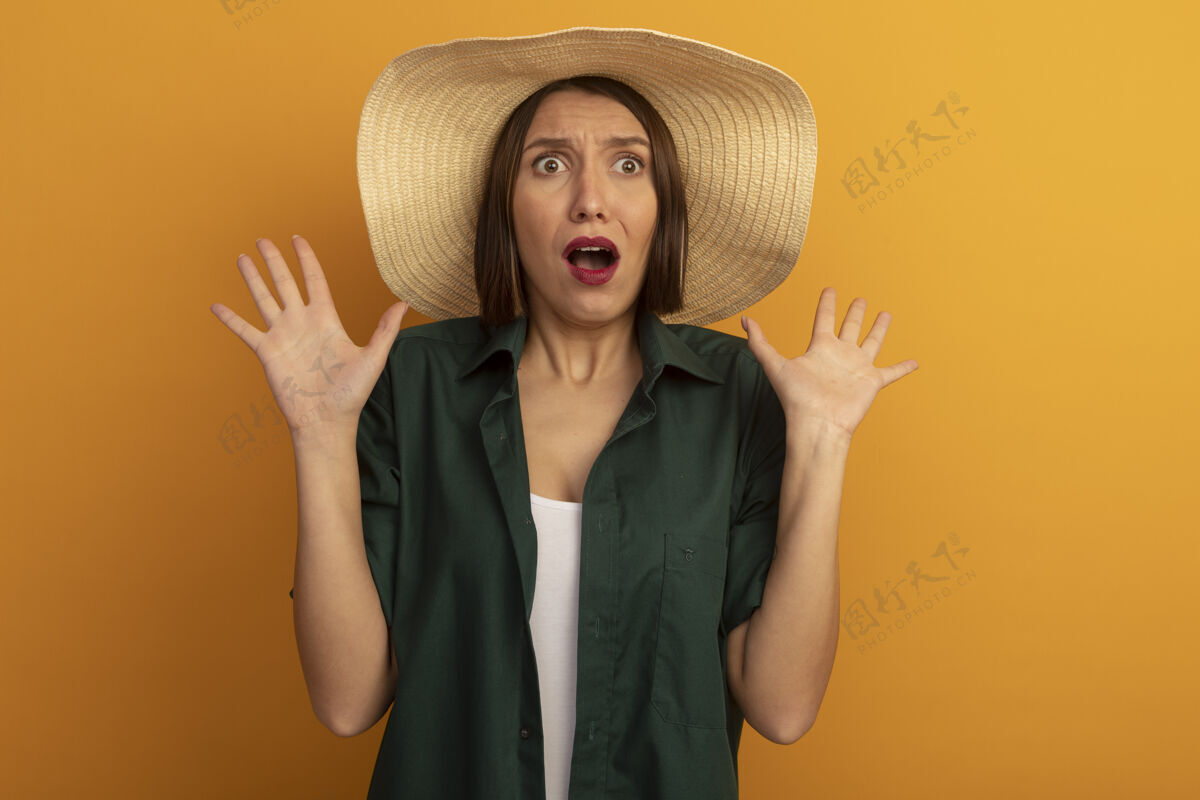 感觉带着沙滩帽的焦急的漂亮女人站在橙色的墙上 举手孤立人姿势人