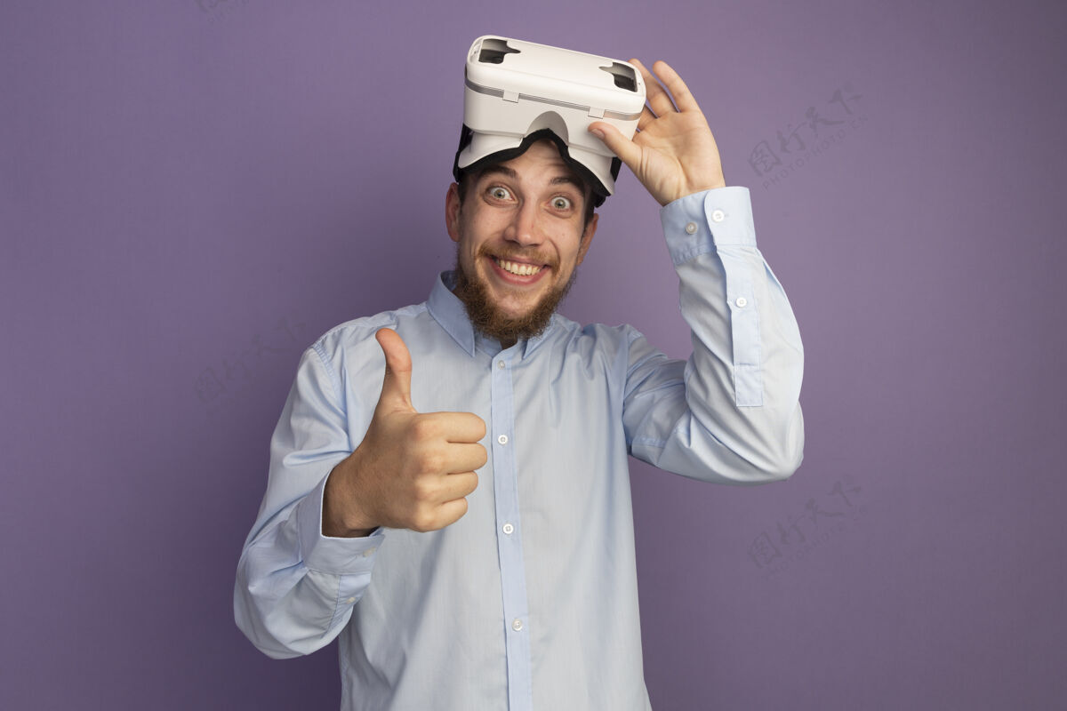 人兴奋的金发帅哥手持vr耳机 竖起大拇指孤立地站在紫色的墙上英俊耳机虚拟现实