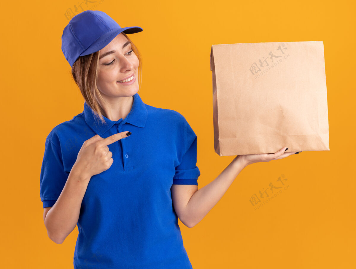 感情身着制服的年轻漂亮的送货员微笑着看着隔离在橙色墙上的纸包装人人漂亮