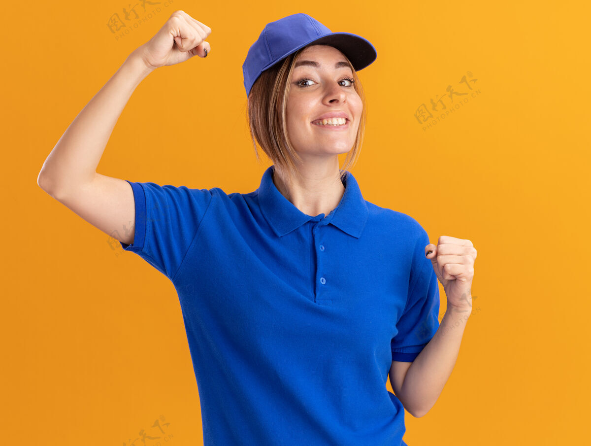 公民微笑着的年轻漂亮的女送货员穿着制服 举起拳头站在橙色的墙上感情制服漂亮