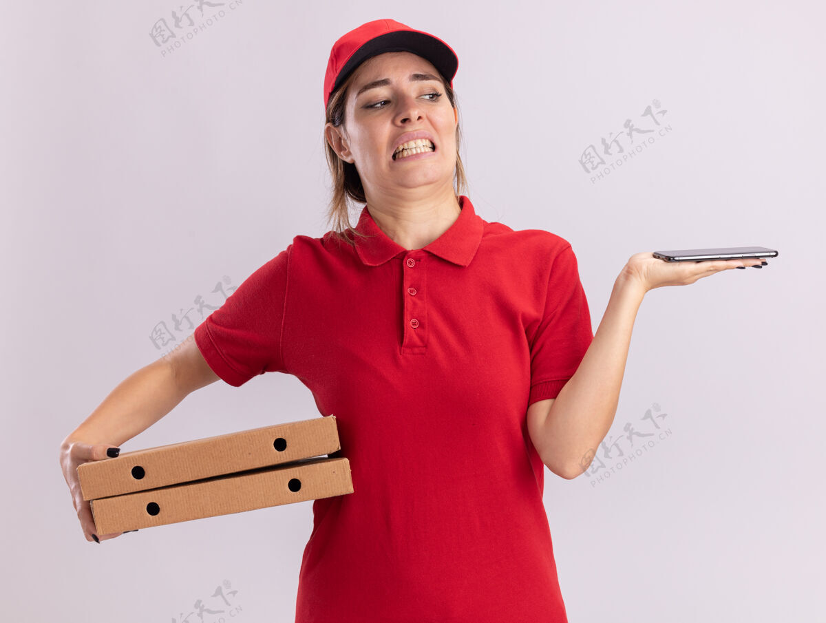 烦恼一个穿着制服的年轻漂亮的送货员手里拿着比萨饼盒 看着隔离在白墙上的电话站着电话姿势