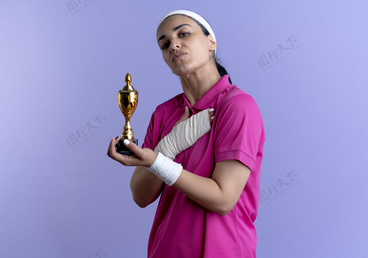 头带年轻自信的白人运动女性戴着头带和腕带 手持冠军杯 将手放在胸部 紫色背景上 留有复制空间佩戴握持手