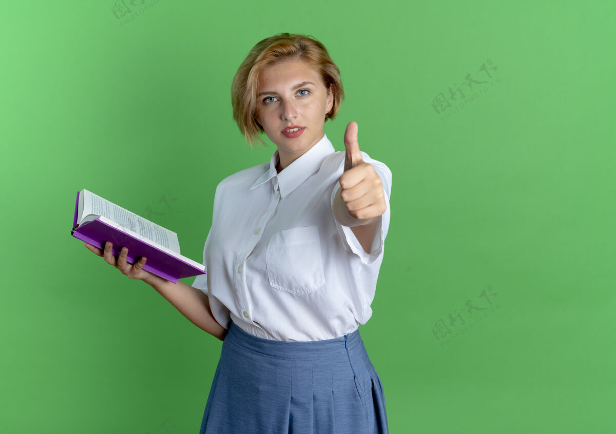 俄国人年轻自信的金发俄罗斯女孩拿着书 竖起大拇指 在绿色的背景上与复制空间隔离年轻坚持金发