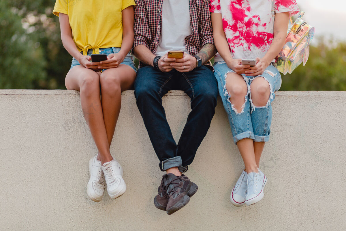 应用程序腿在运动鞋的年轻朋友公司坐在公园使用智能手机学生无线组