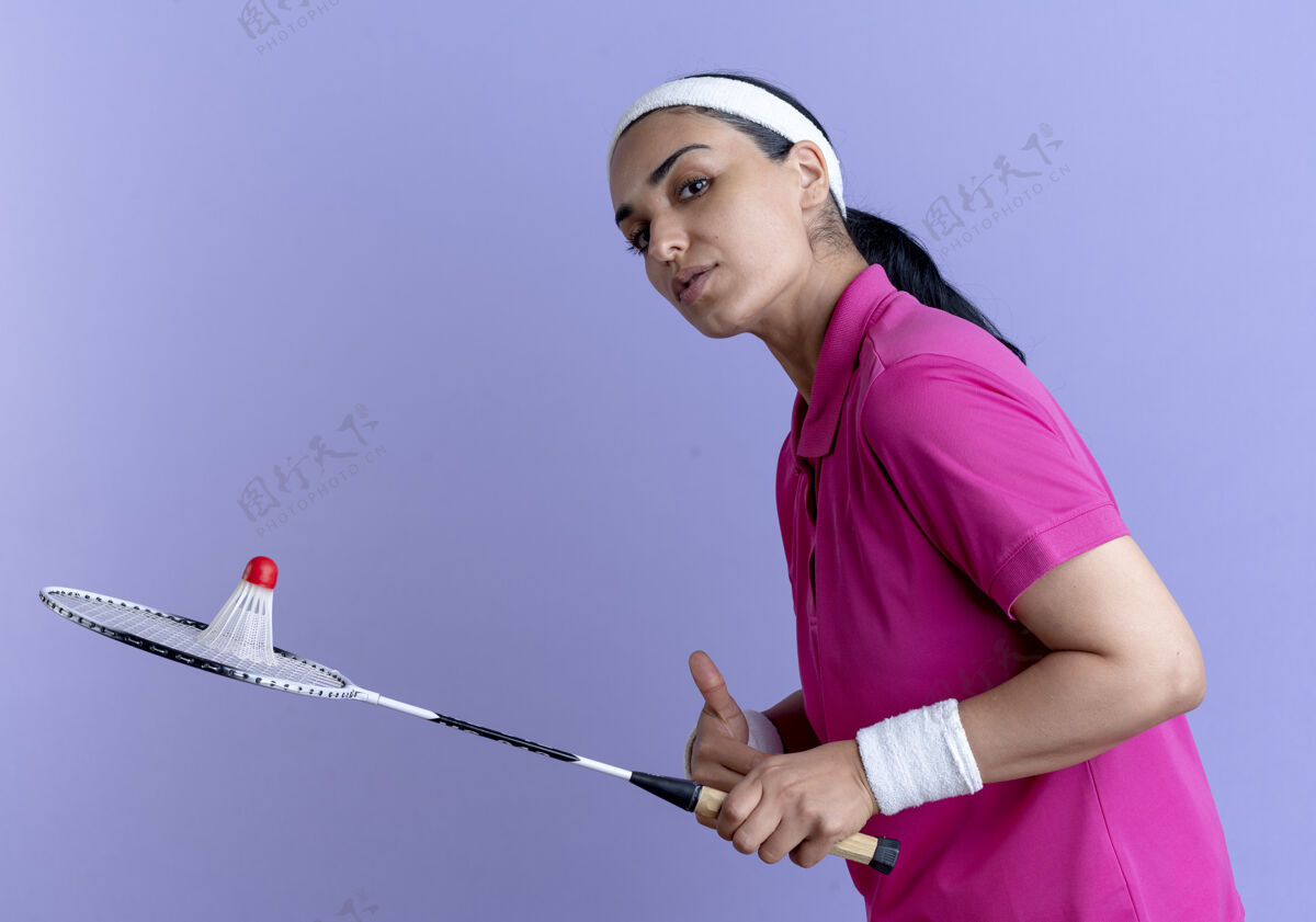 头带年轻自信的白人运动女性戴着头带和腕带站在一边拿着羽毛球在球拍上紫色背景与复制空间隔离穿自信立场