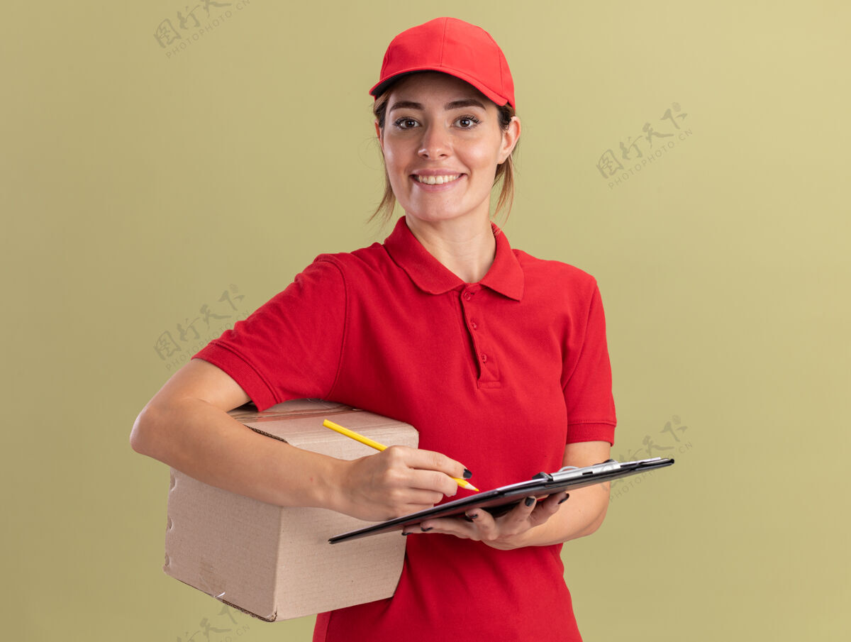 立场微笑着的年轻漂亮的女送货员穿着制服 用铅笔夹着卡片盒写在剪贴板上 隔离在橄榄绿的墙上年轻脸姿势