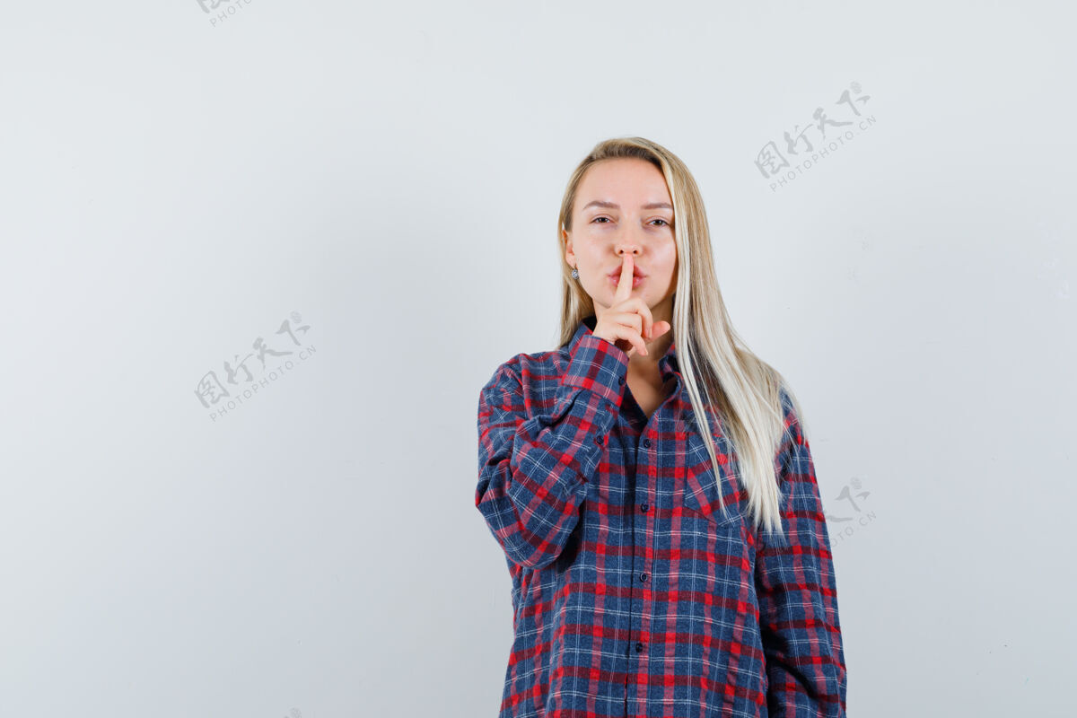 显示金发女人把食指放在嘴上 穿着格子衬衫 表现出沉默的姿态 看起来很有吸引力 正面视图嘴巴衬衫漂亮