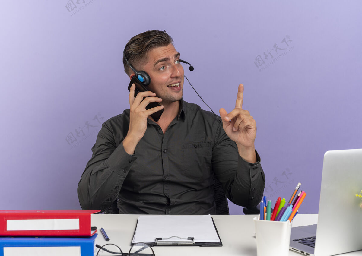 惊喜年轻的金发上班族戴着耳机坐在办公桌旁 手里拿着办公工具 用手提电脑在电话里讲话 指着紫色背景上孤立的 有复印空间的地方金发坐着指着