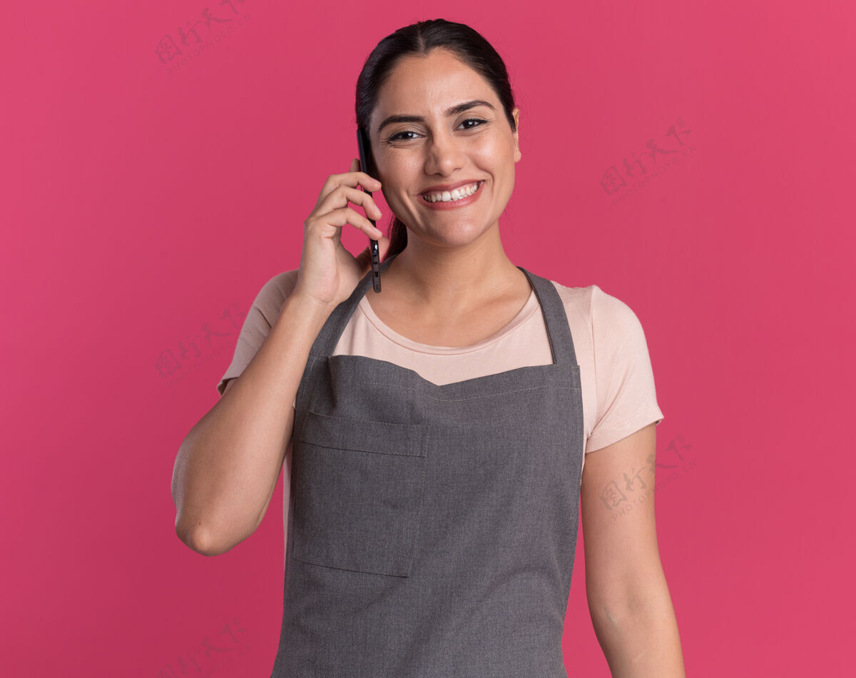 快乐站在粉红色的墙上 年轻漂亮的女理发师围着围裙 面带微笑地看着前面 一边讲着手机漂亮手机围裙