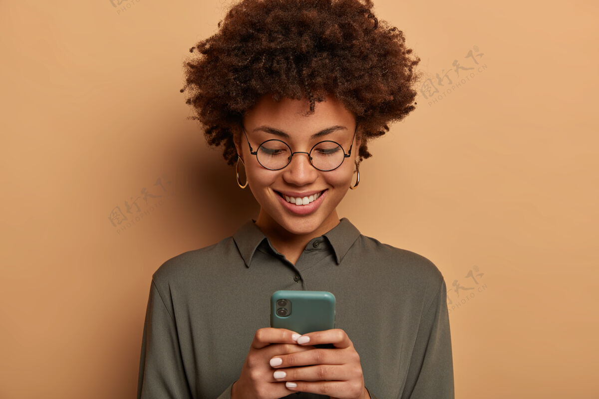 眼镜卷发女士头戴透明眼镜 灰色衬衫 在智能手机上使用免费在线应用程序 查看图像 戴圆眼镜和衬衫快乐皮肤用户
