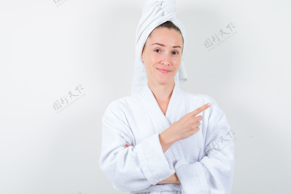 头发年轻女士指着右上角的白色浴衣 毛巾 看起来很高兴前视图模特人女性