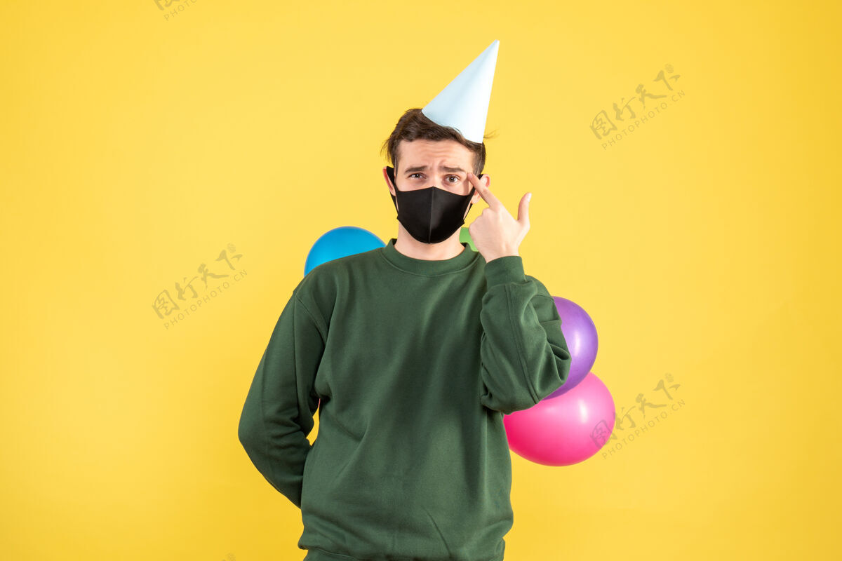 人物正面图：戴着派对帽 站在黄色背景上的彩色气球的年轻人前面面具剪影