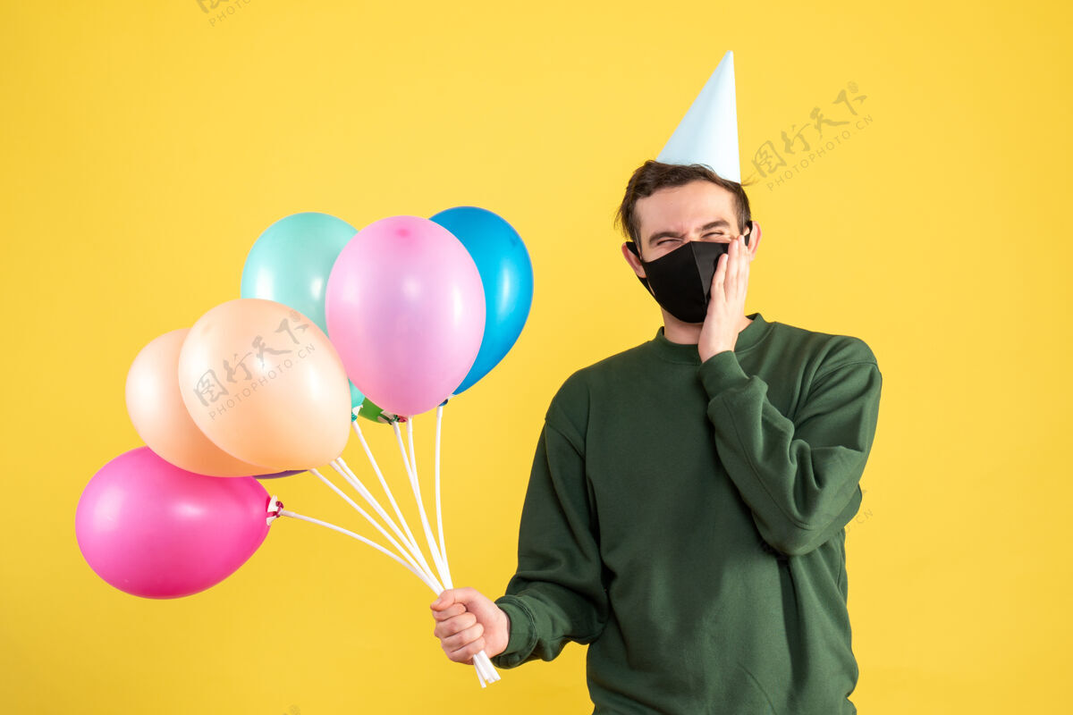 帽子正面图：戴着派对帽 站在黄色背景上的彩色气球的年轻人生日背景气球