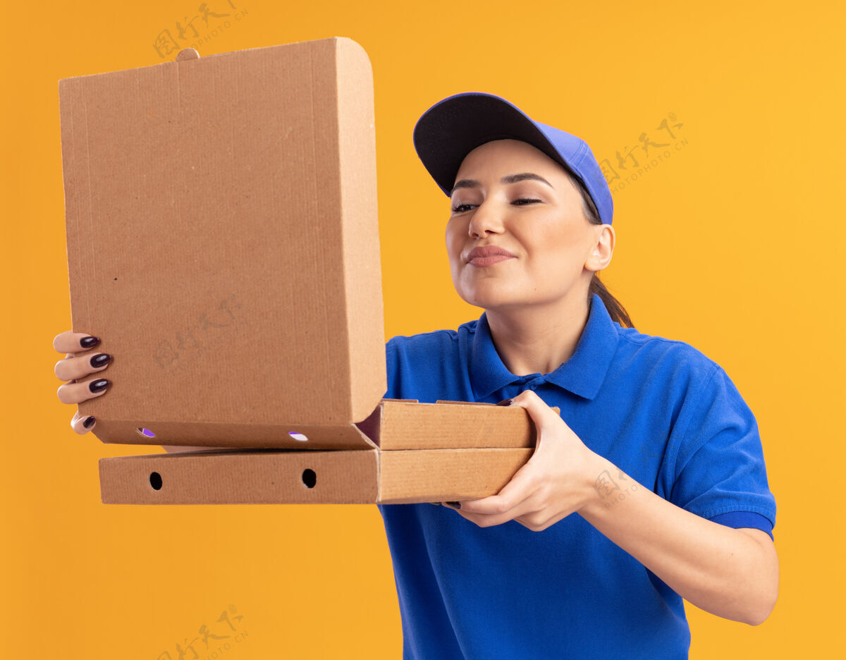 站着身着蓝色制服 戴着帽子的年轻送货员站在橘色的墙上 手里拿着披萨盒 打开盒子 吸入着怡人的香气拿着香气开着