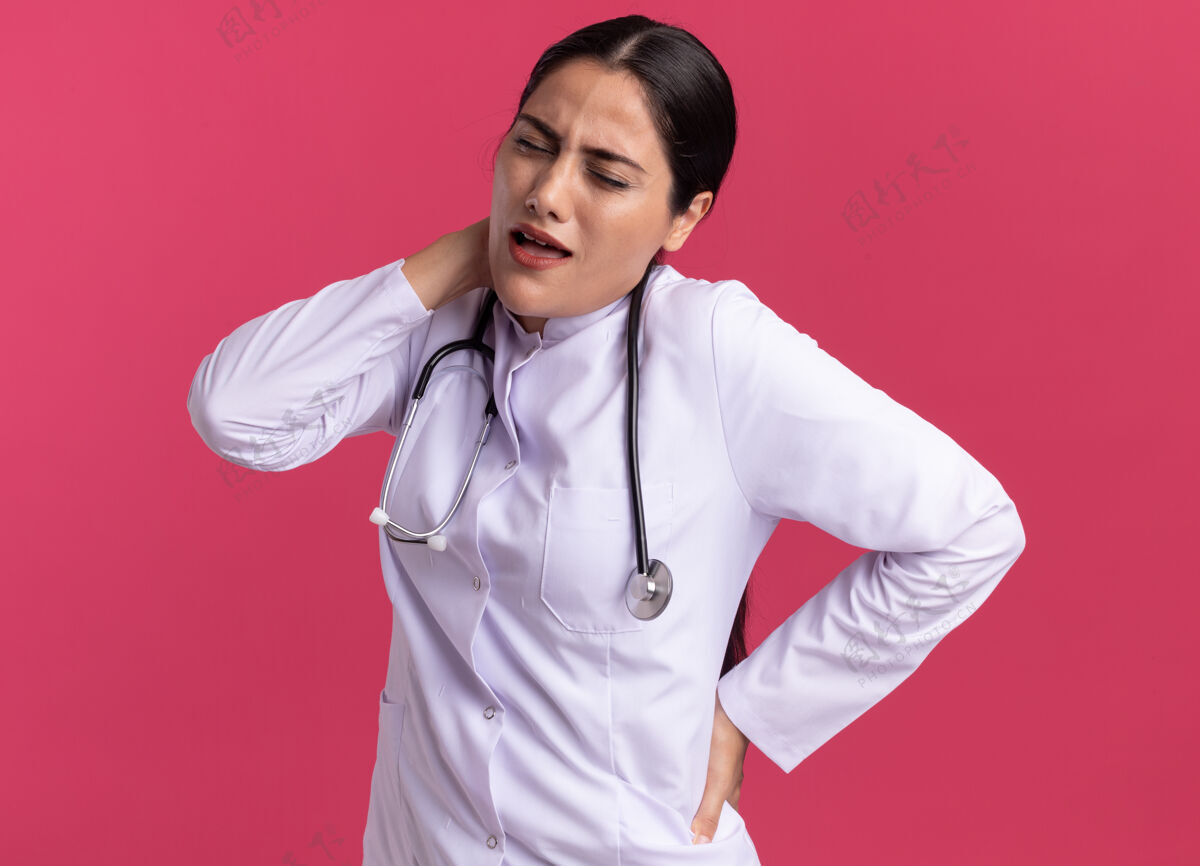 外套穿着医用外套的年轻女医生 戴着听诊器 看着不舒服 摸着脖子 站在粉红色的墙上感到疼痛医生听诊器站