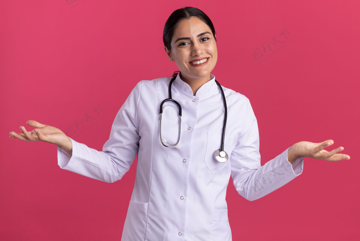 站立身穿医用外套 手持听诊器的年轻女医生站在粉红色的墙上 面带微笑 高举双臂 看着前方医生医生女人
