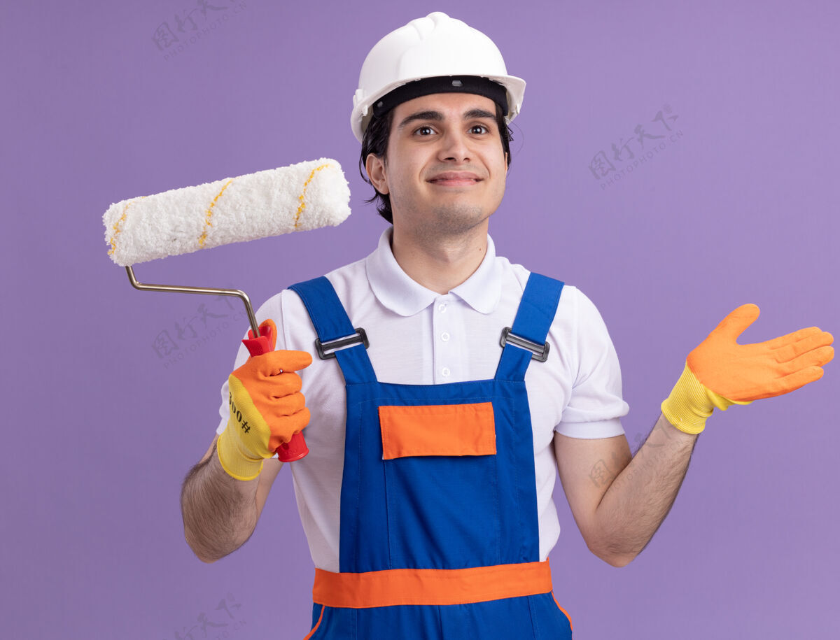 手套年轻的建筑工人穿着建筑制服 戴着安全帽 戴着橡胶手套 拿着油漆辊 面带微笑地站在紫色的墙上 望向一边站安全微笑