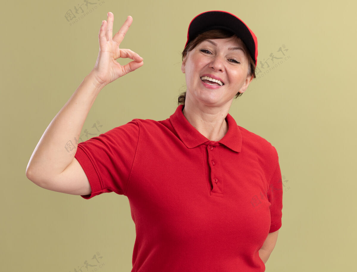 帽子身穿红色制服 头戴帽子的中年女送货员站在绿色的墙上 面带微笑 脸上挂着“ok”的牌子好中间年龄
