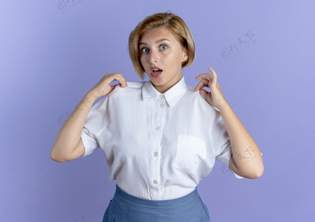 惊喜年轻惊讶的金发俄罗斯女孩拿着衬衫看着相机在紫色背景上与复制空间隔离拿着相机年轻