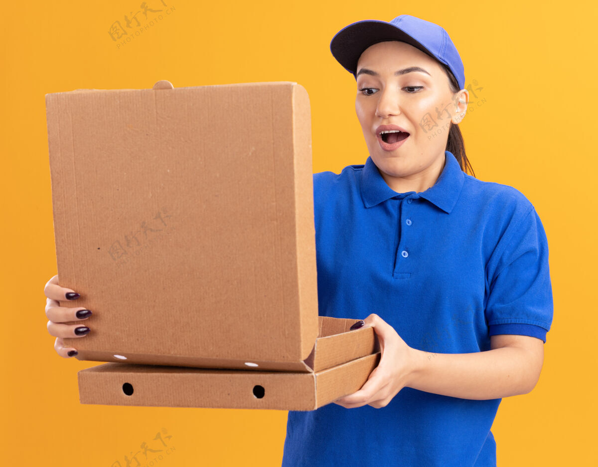 拿着身穿蓝色制服 头戴鸭舌帽的年轻女送货员拿着披萨盒打开盒子 站在橙色的墙上惊讶地看着它制服太棒了开着