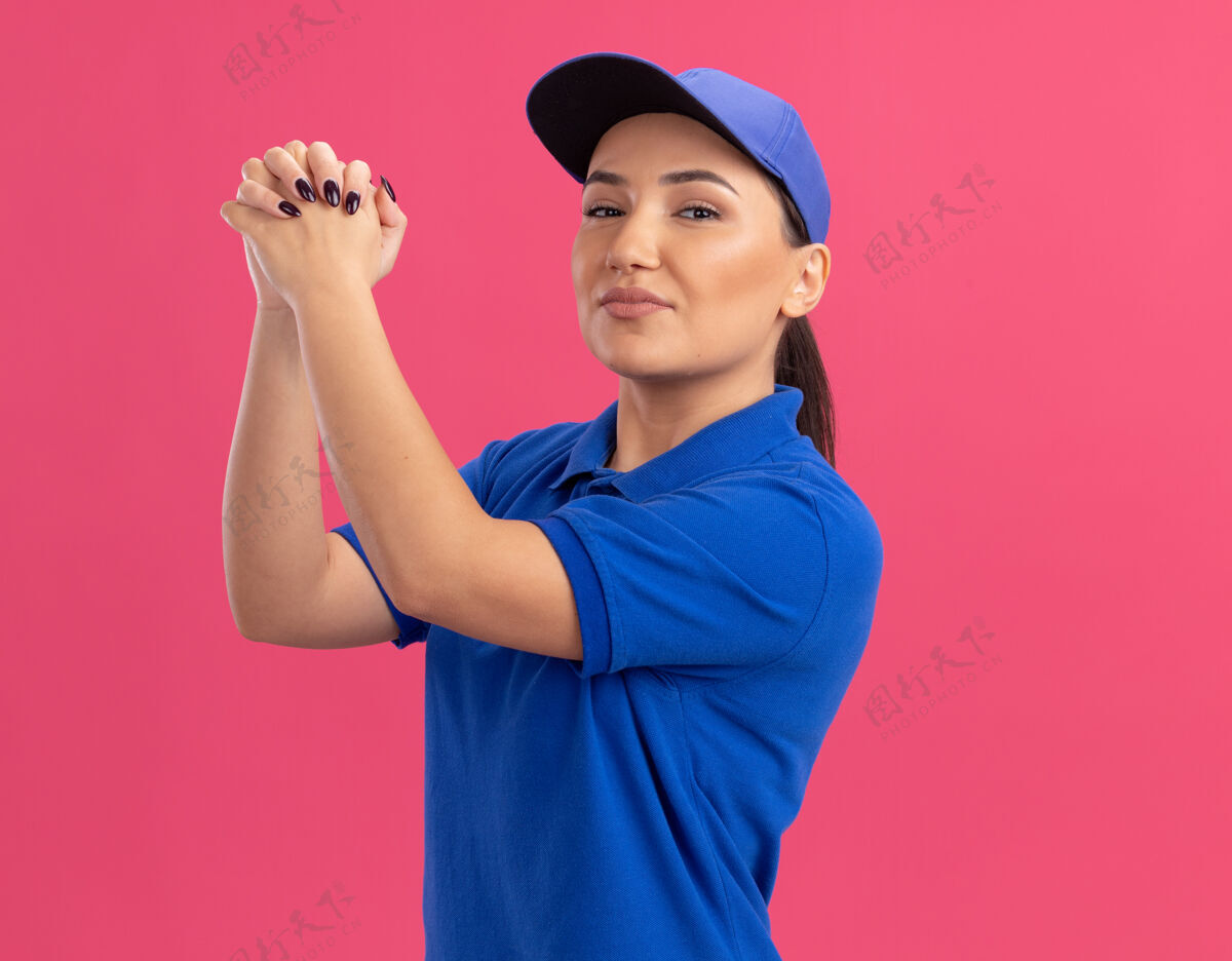 手势身穿蓝色制服 头戴鸭舌帽的年轻送货员站在粉红色的墙上 微笑着自信地做着团队合作的手势微笑女人信心