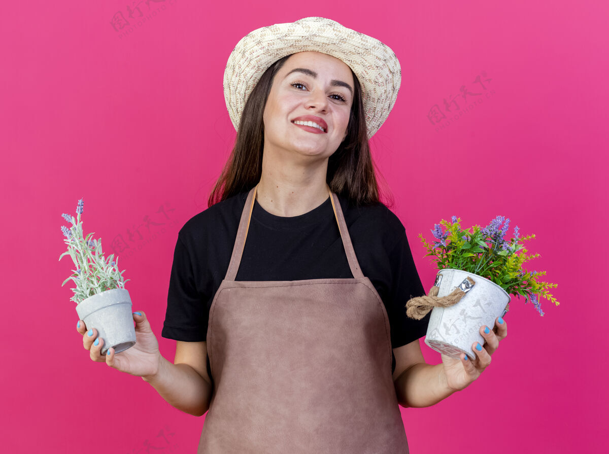 举行微笑美丽的园丁女孩穿着制服 戴着园艺帽 在粉红色背景上的花盆里捧着花帽子女孩花盆